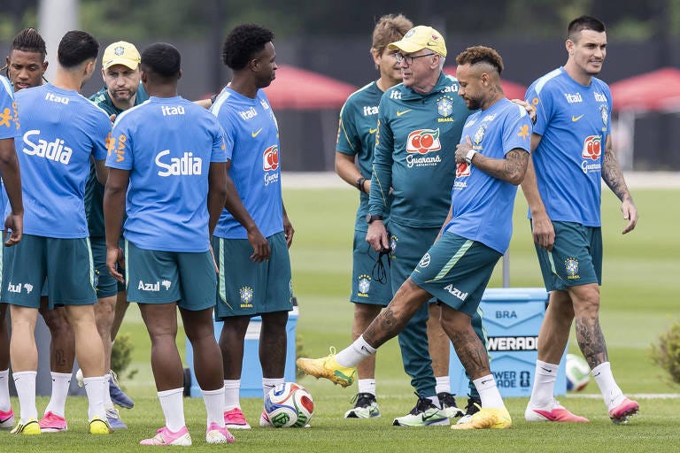 Jogadores da seleção brasileira treinam em campo aberto, com técnico observando próximo. Um jogador chuta a bola enquanto outros aguardam ao redor, todos usando uniformes azuis e verdes.