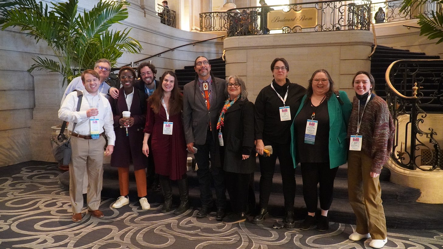 Ten adults stand with arms interlaced in a hotel lobby with a sizable curving set of steps behind them. They’re wearing conference lanyards and smiling at the camera. It is daytime. Ten adults stand with arms interlaced in a hotel lobby with a sizable curving set of steps behind them. They’re wearing conference lanyards and smiling at the camera. It is daytime.