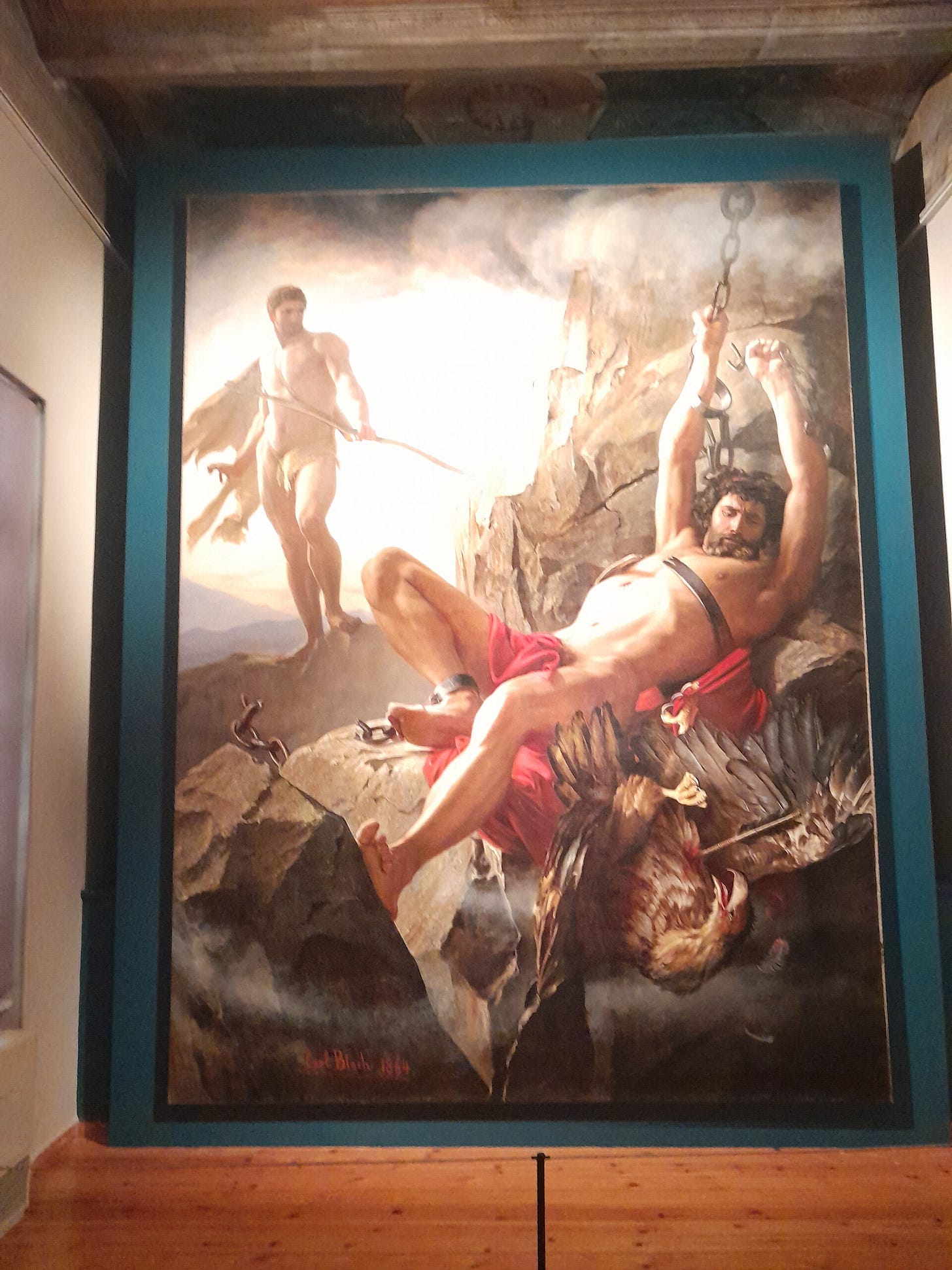 A huge vertical canvas on a gallery wall: a muscular man in a red cloth hangs from chains on a rock, one arm still shackled, while a dead eagle with an arrow through it falls at his feet and Heracles, holding a bow, watches from the left.