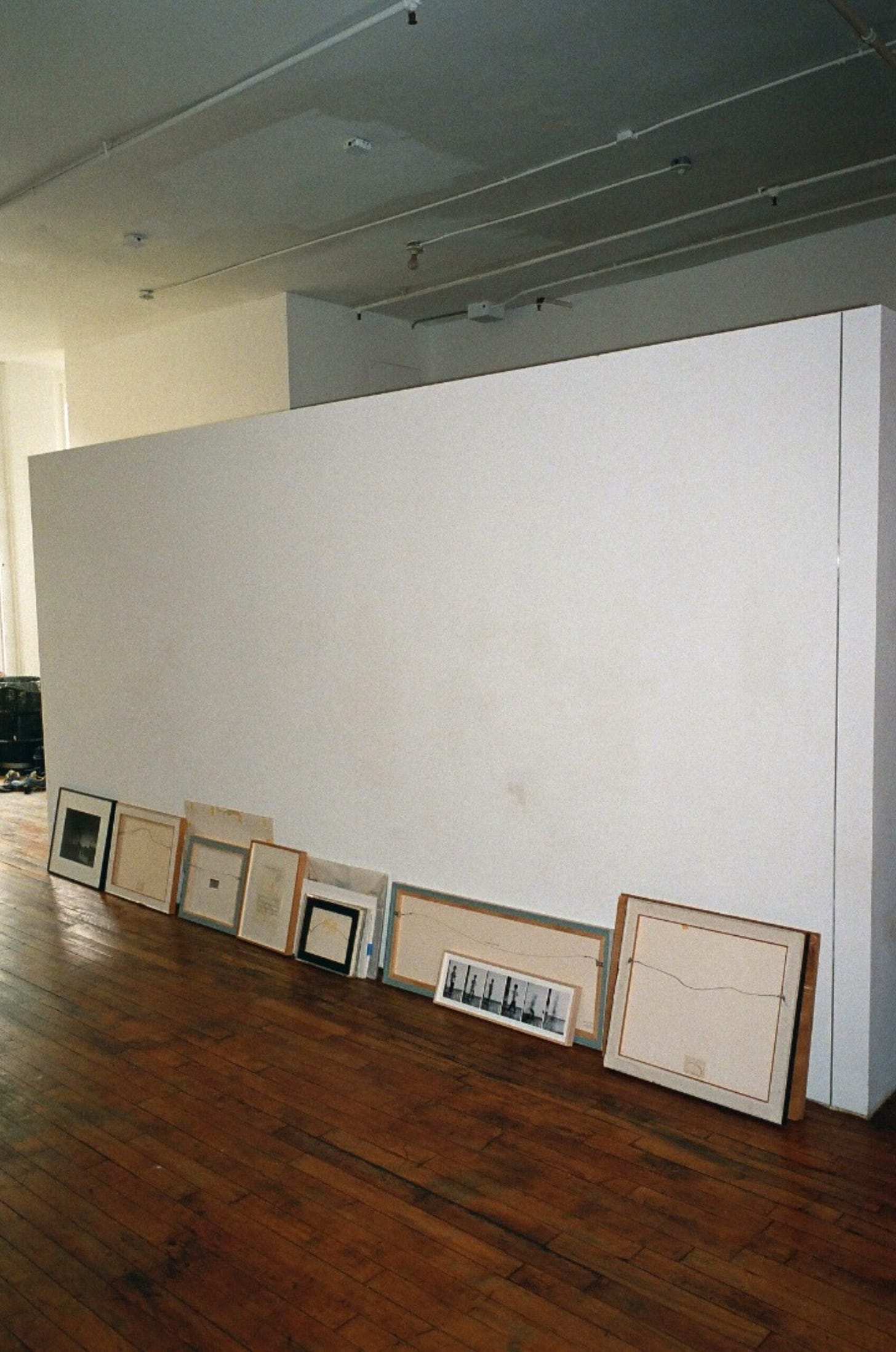 Framed artworks turned around and leaning against a gallery wall along a wood floor.