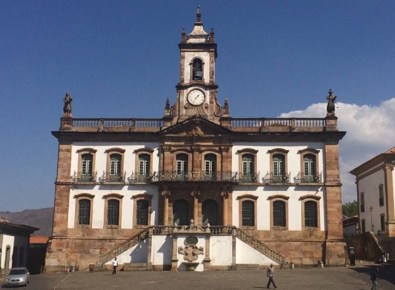 #descrevemospravocê  A imagem mostra a fachada do Museu da Inconfidência, localizado em Ouro Preto, Minas Gerais. O prédio histórico é imponente, com três pavimentos e arquitetura colonial barroca. A construção é simétrica e feita em alvenaria branca com detalhes em pedra sabão. No centro da fachada, há uma escadaria dupla que se une em um patamar elevado diante da entrada principal. Acima da entrada, está uma varanda com grades de ferro trabalhado. No segundo andar, vê-se uma fileira de janelas com molduras de pedra, todas com venezianas verdes fechadas. Na parte superior central do edifício, destaca-se uma torre com relógio redondo e sino, encimada por uma pequena estrutura em forma de campanário. No topo do prédio, quatro esculturas humanas decoram os cantos da balaustrada. O céu ao fundo está limpo, com poucas nuvens e luz solar intensa, o que projeta sombras nítidas na fachada. Algumas pessoas caminham na praça em frente ao prédio, e um carro branco está estacionado à esquerda.
