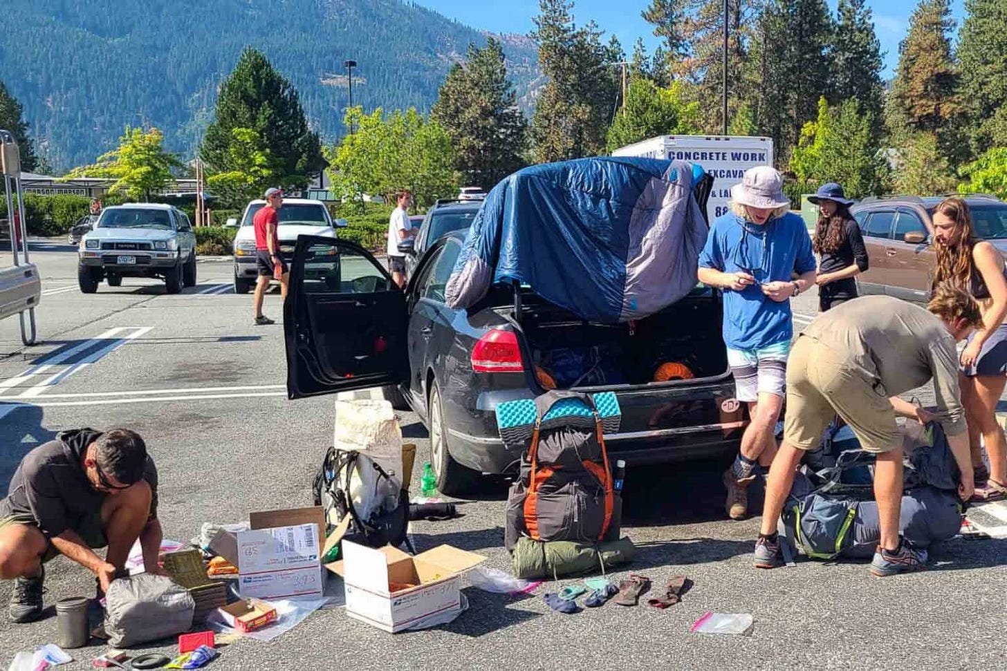A group of thru-hikers unpack and organize resupply boxes in a parking lot, surrounded by open backpacks, gear, and mail parcels. A car trunk is open with gear spilling out, and a sleeping bag is draped over the back window. Mountains and trees rise in the background on a sunny day.