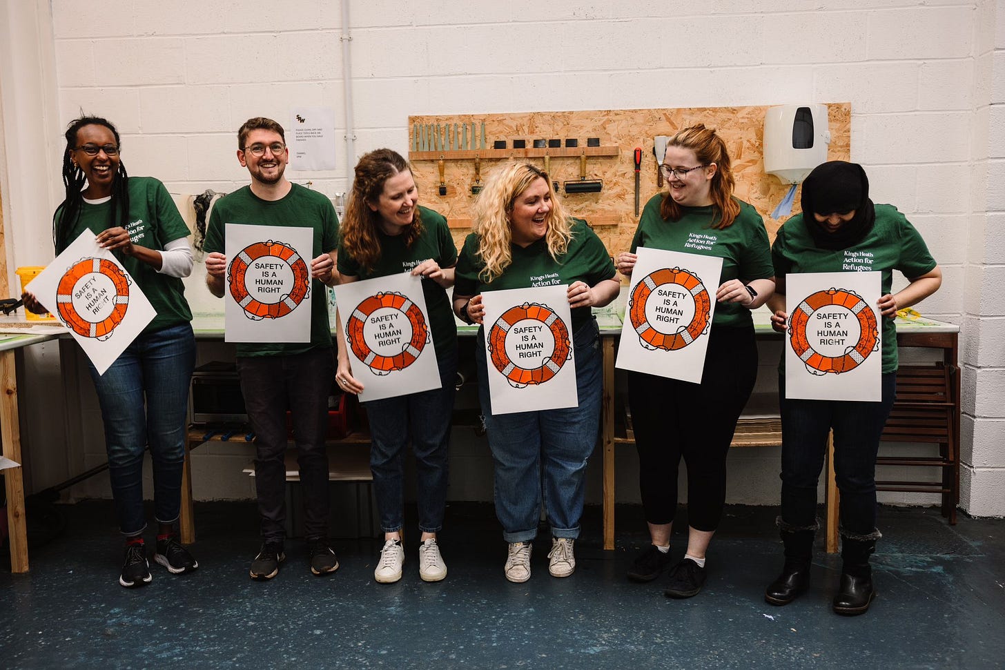 Image of six Kings Heath Action for Refugees volunteers in matching green tshirts, holding up artwork featuring orange dinghys with text 'safety is a human right'. The six are laughing and it is a candid photo.