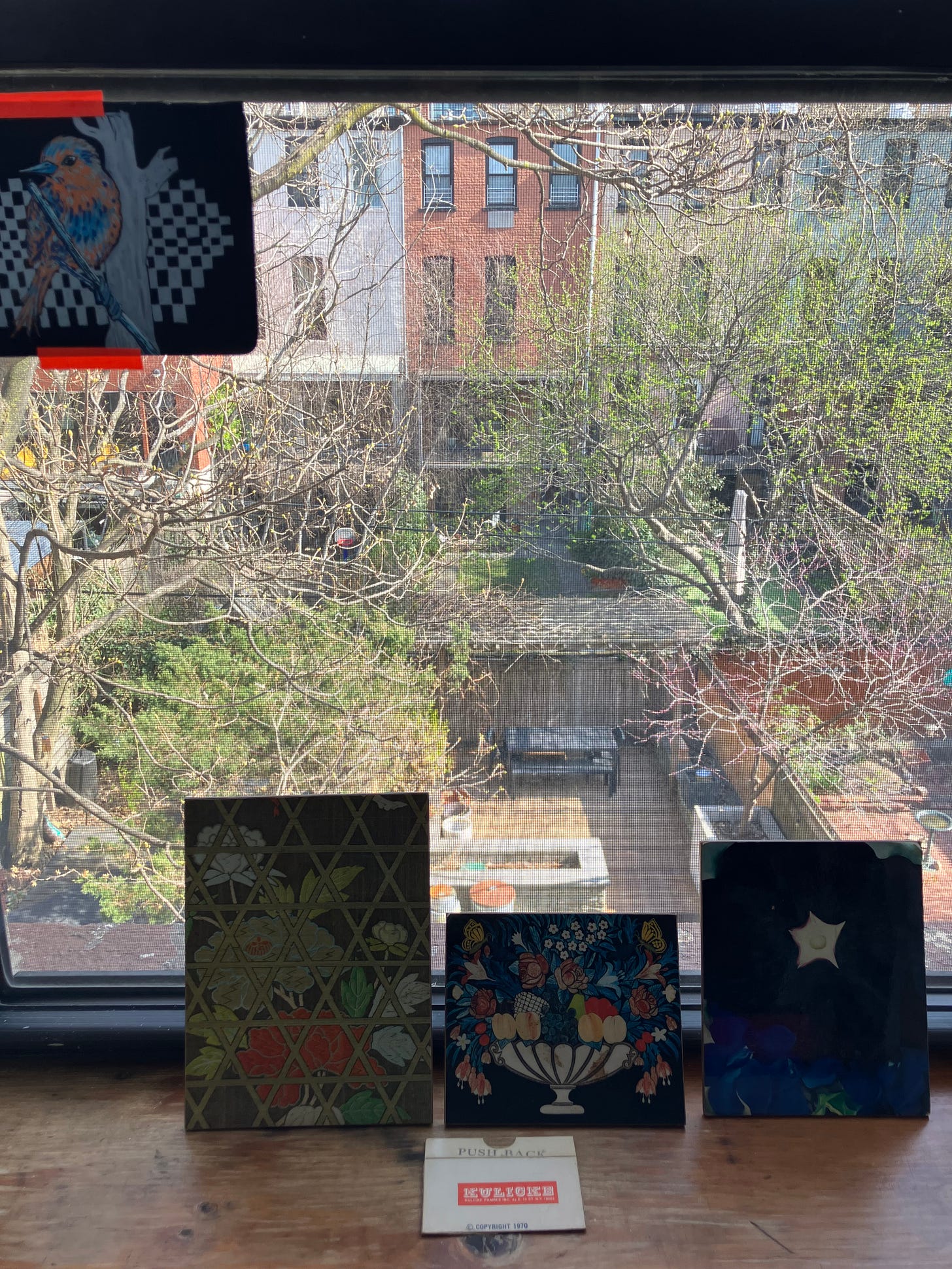 view of window with three floral pictures on sill and drawing of orange and blue bird taped to glass. Trees and back yard visible below.