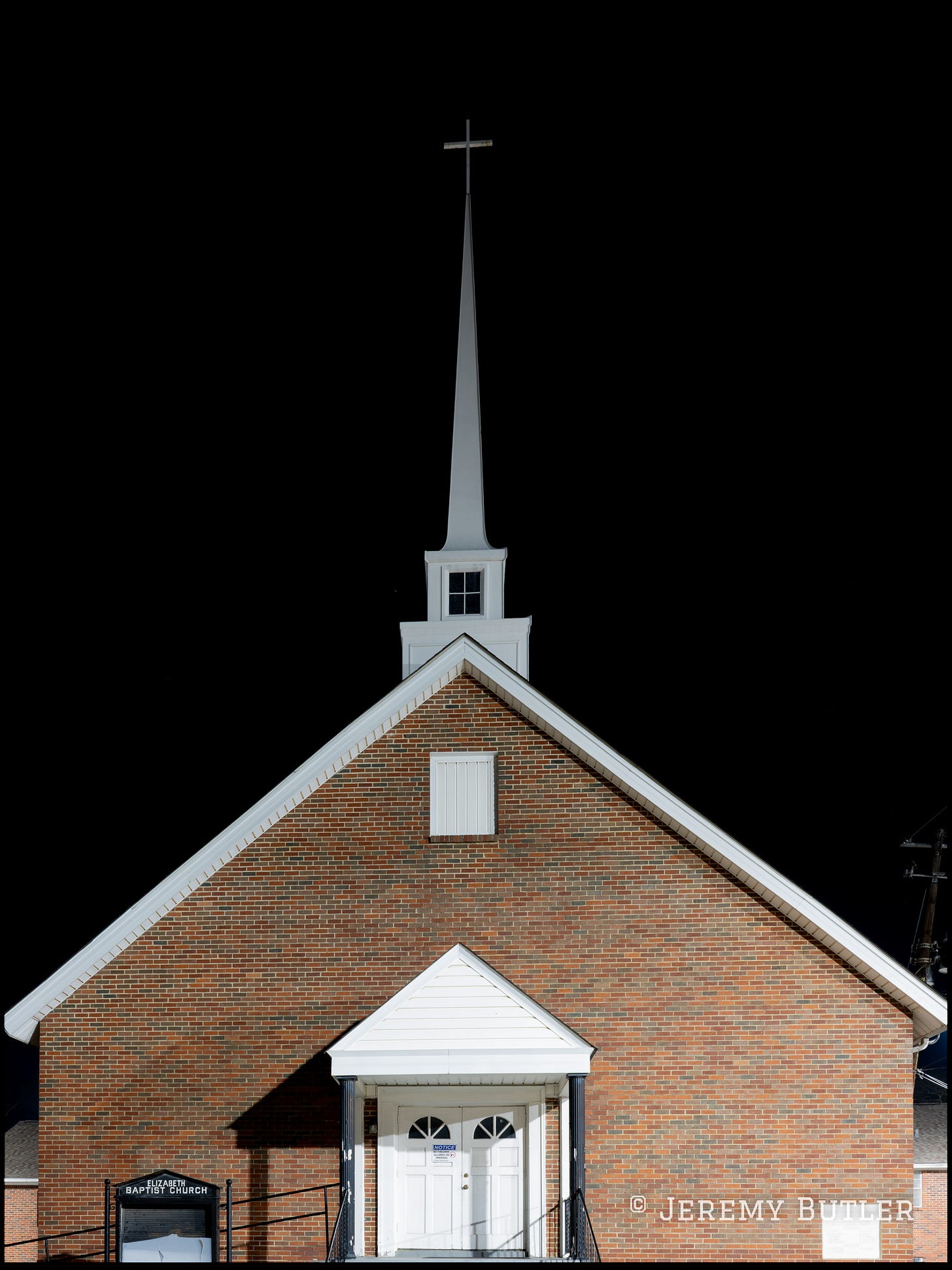 A red-brick Baptist church with a tall white steeple and cross photographed at night.