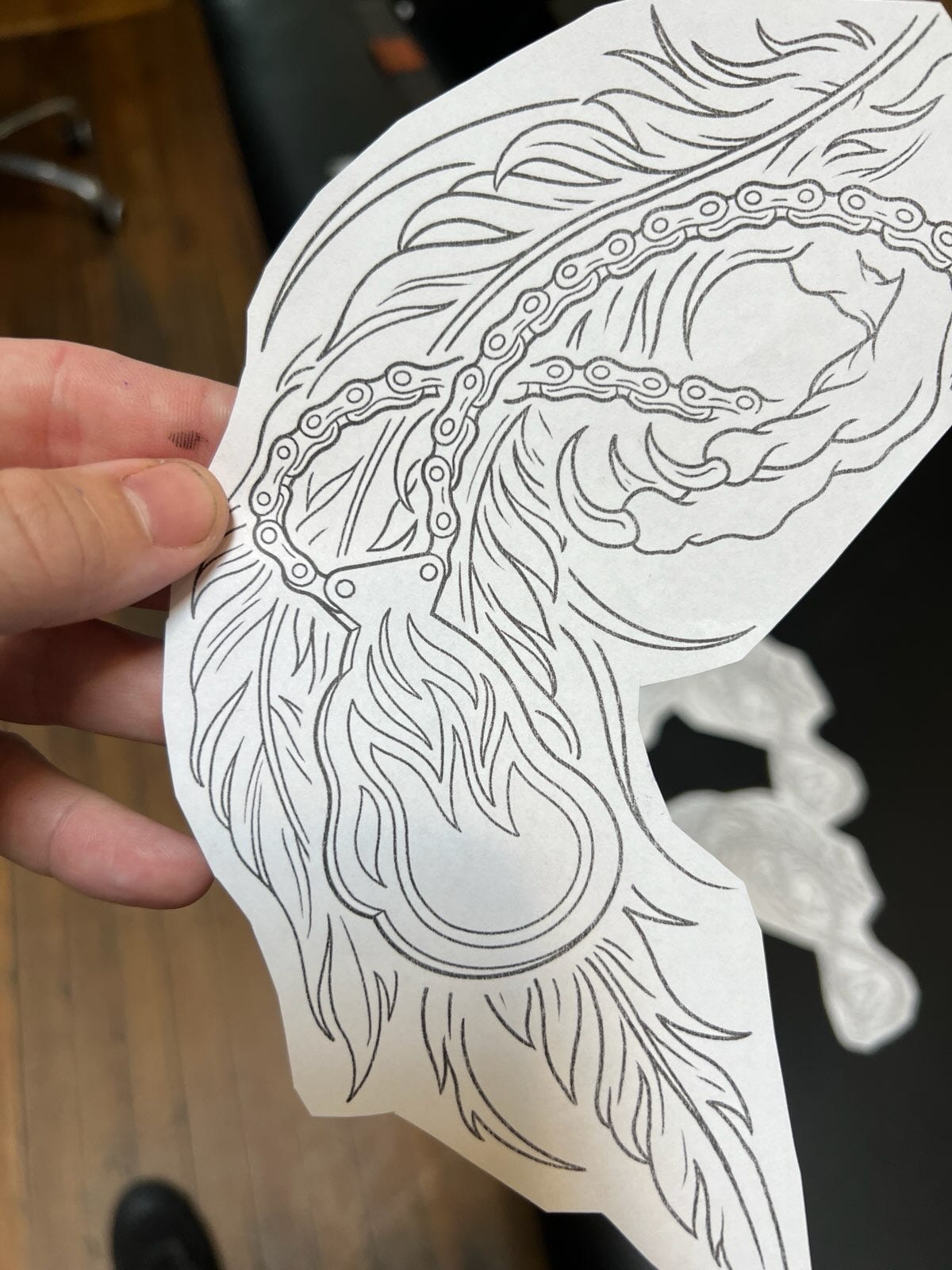 A photograph of a hand holding a cutout of a tattoo stencil. It is a black-and-white line drawing showing the claws of a phoenix holding a bicycle chain. There is a flame-shaped medallion connected to the chain as a link. In the background are flames of the firebird’s tail. A photograph of a hand holding a cutout of a tattoo stencil. It is a black-and-white line drawing showing the claws of a phoenix holding a bicycle chain. There is a flame-shaped medallion connected to the chain as a link. In the background are flames of the firebird’s tail.