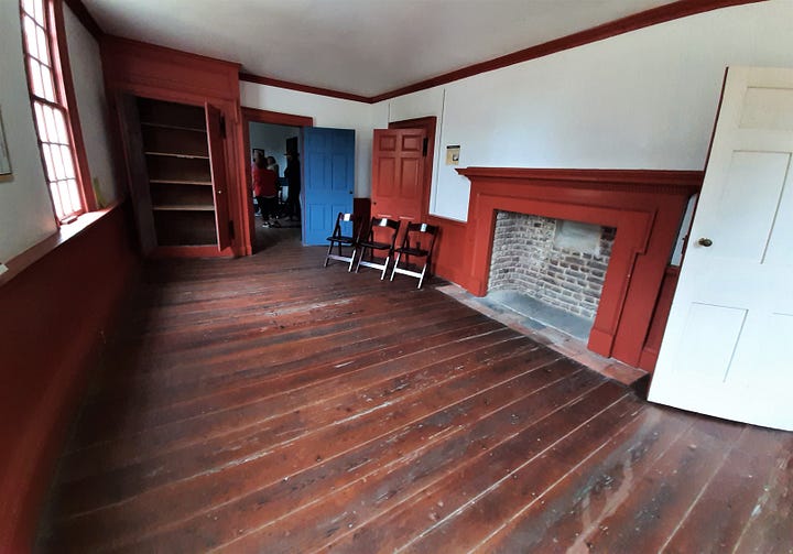 The interior of Hampton shows large rooms with wooden floors. The paint is blue. The fireplace is brick, with chairs sitting around.