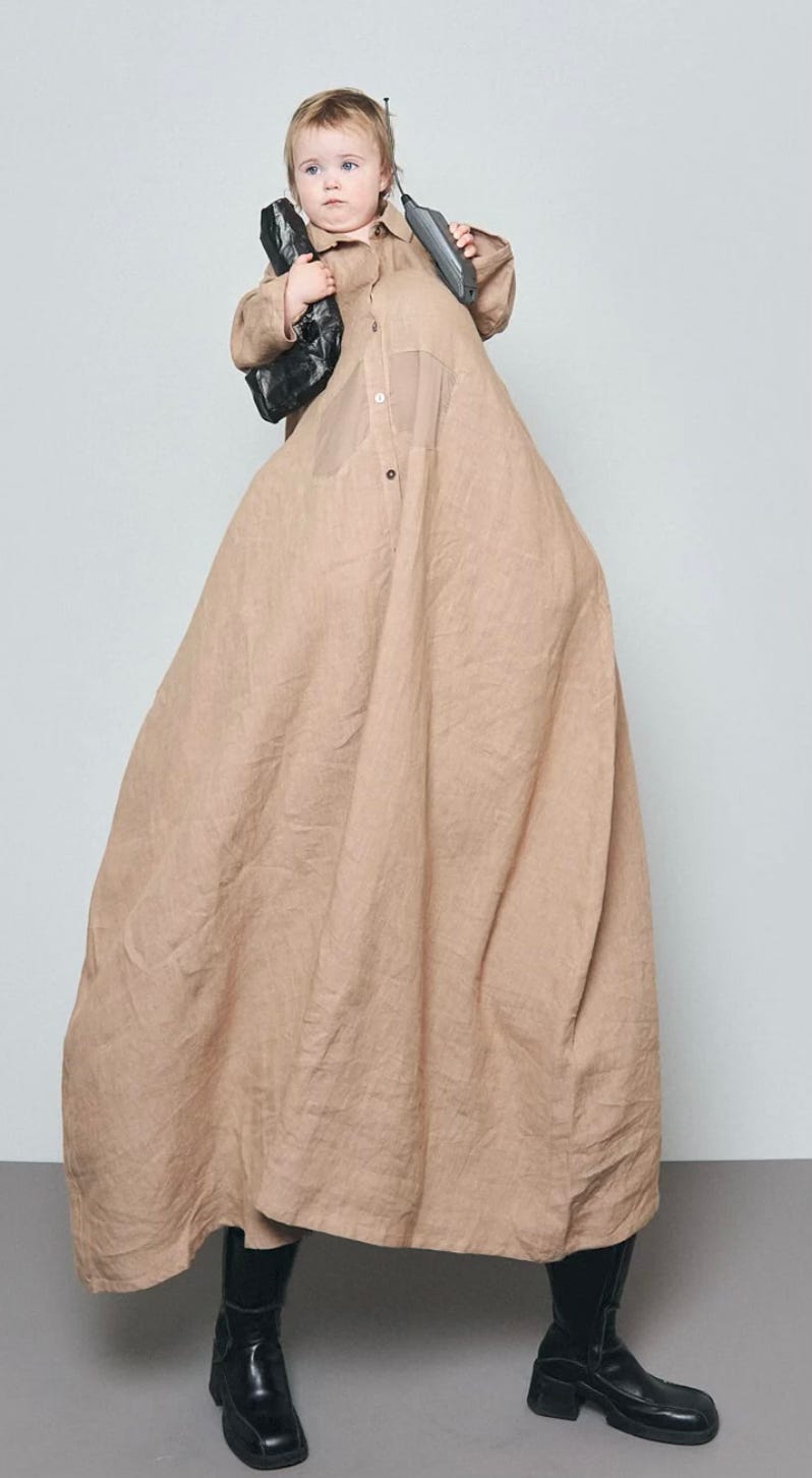 a toddler holding a 90s style chunky cell phone in one hand and an unidentified bag in the other while wearing a very very long tan trench coat. The coat is so long that it’s clear there is a person under it holding the child on their shoulders. The toddler looks like he has absolutely no idea what is going on.