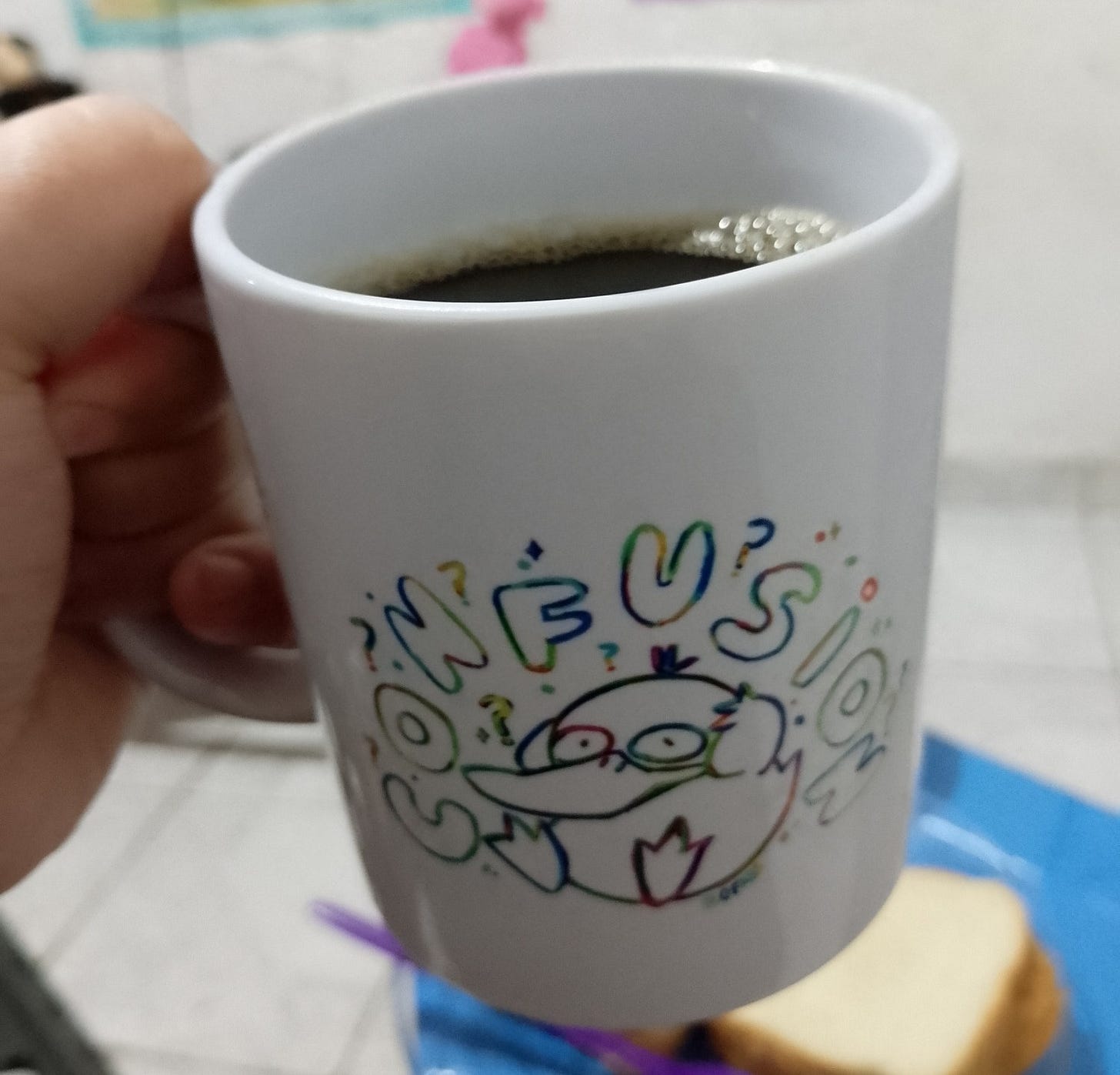 A imagem mostra uma caneca branca com café, nela há uma imagem do Pokemon Psyduck e a palavra "CONFUSION" escrita de forma estilizada acima dele.