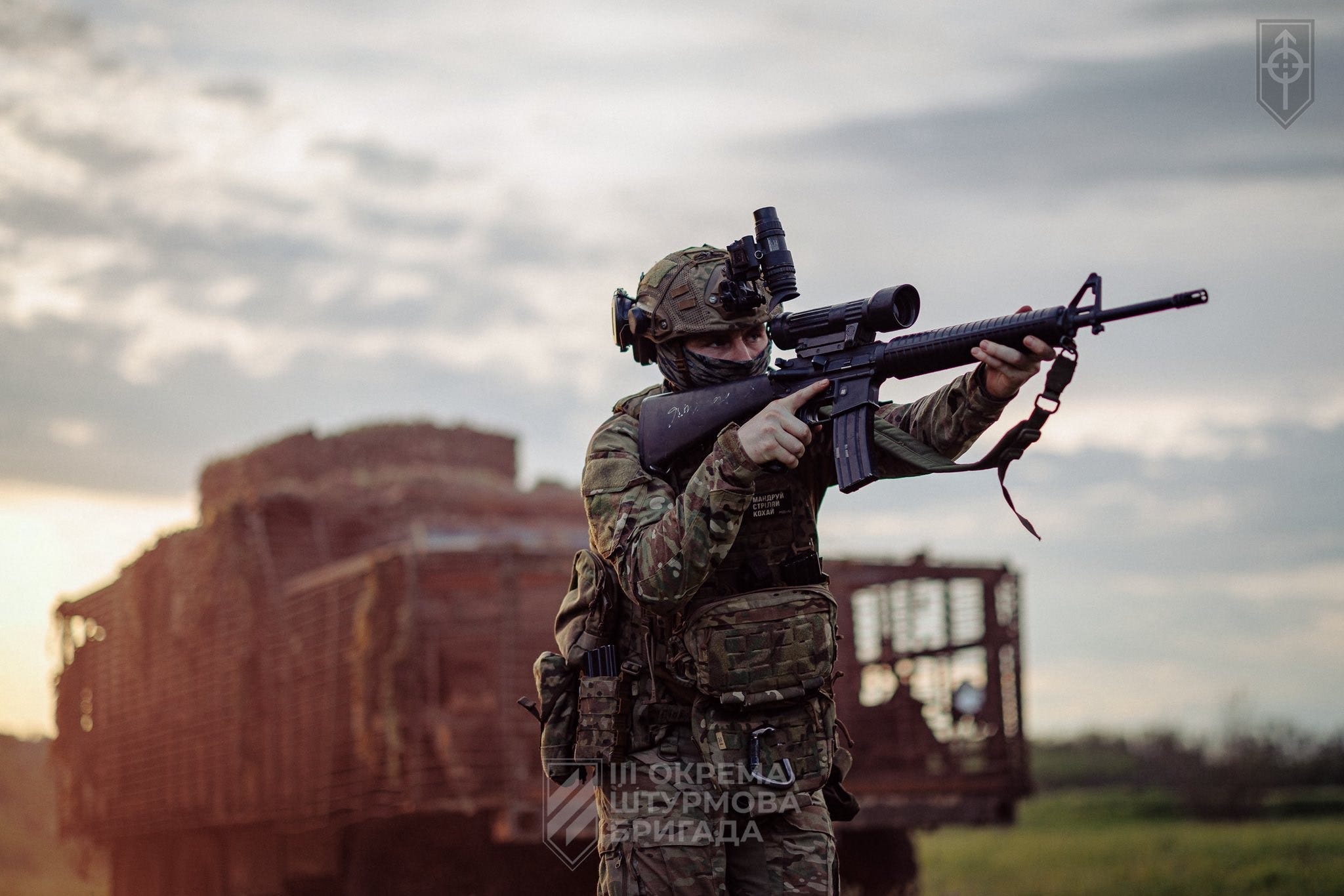 A Ukrainian soldier in a helmet with night-vision mount and multicam gear aims a scoped rifle skyward in an open field at dusk, with a camouflaged truck behind him. A Ukrainian soldier in a helmet with night-vision mount and multicam gear aims a scoped rifle skyward in an open field at dusk, with a camouflaged truck behind him.