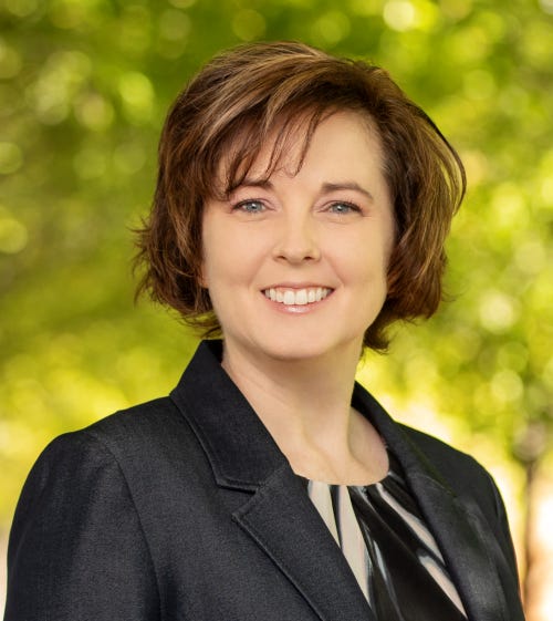 A professional headshot of a woman with short brown hair and blue eyes, smiling warmly. She is wearing a dark blazer over a black-and-white patterned blouse. The background is softly blurred with bright green foliage, suggesting a natural outdoor setting in daylight.