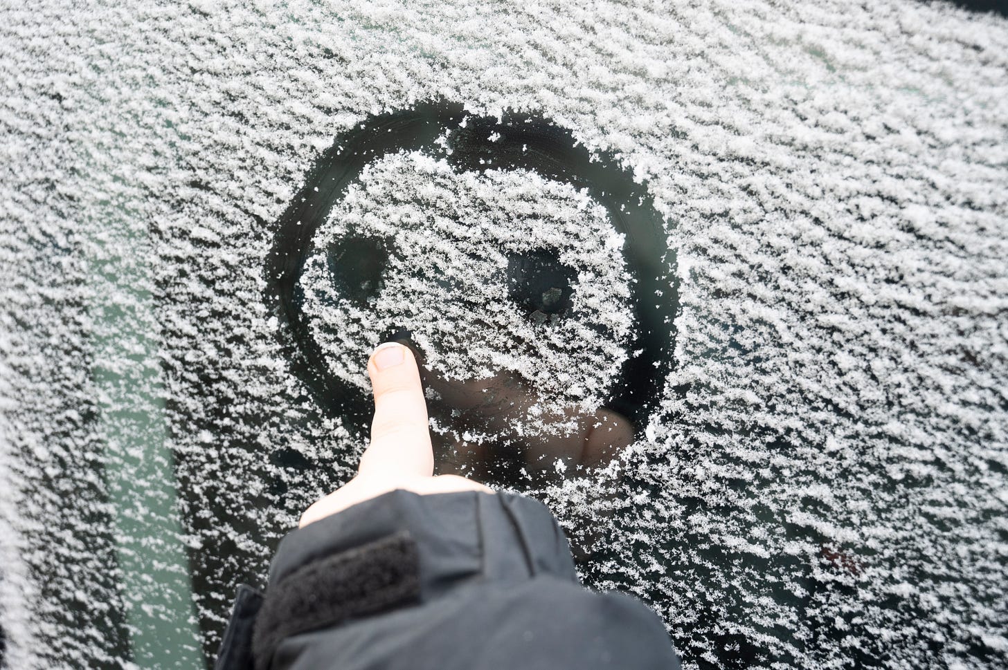 A middle finger poking out of a black coat sleeve drawing a smiley face on a snowy window