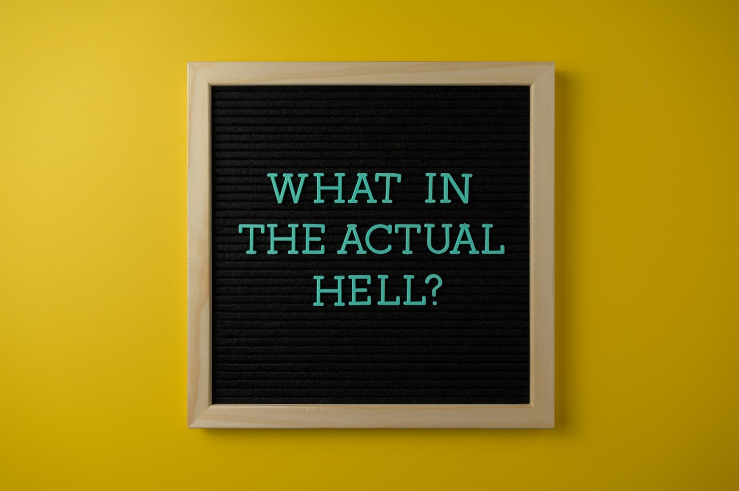a blackboard with the words what in the actual hell written in teal - the board sits on a yellow wall