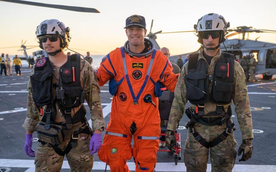 A smiling astronaut in an orange spaceflight suit is escorted on the deck of a ship by two men in military gear. A smiling astronaut in an orange spaceflight suit is escorted on the deck of a ship by two men in military gear.