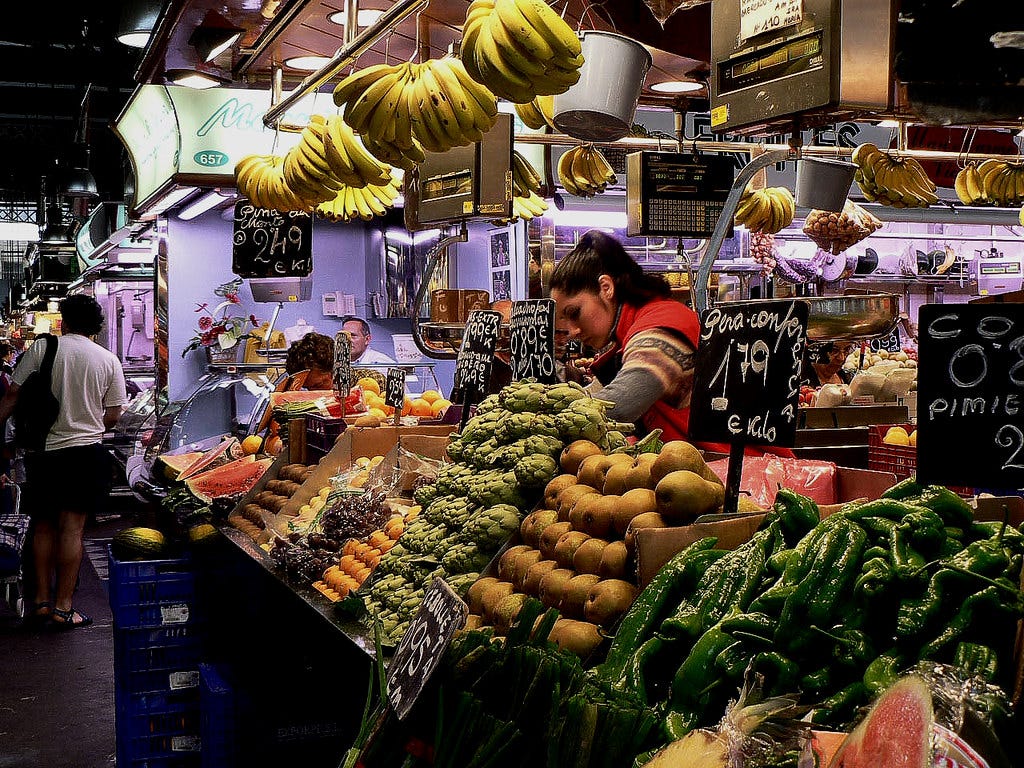 Fruits and vegetables are stacked in colorful rows at a produce stall in an enclosed market hall. A woman bends over the produce near the counter. Fruits and vegetables are stacked in colorful rows at a produce stall in an enclosed market hall. A woman bends over the produce near the counter.