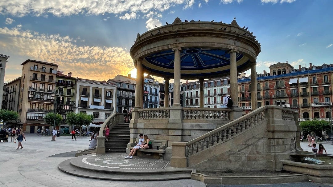 A square surrounded by high, colorful houses. Central to the photo is a round stone bandstand. The orange sky indicates an early sunset.