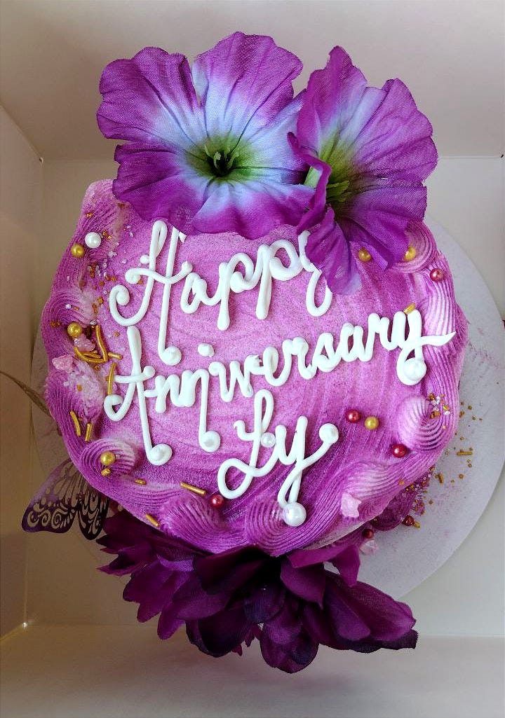 A cake decorated with purple frosting and purple flowers. Says "Happy Anniversary Ly" in white frosting.