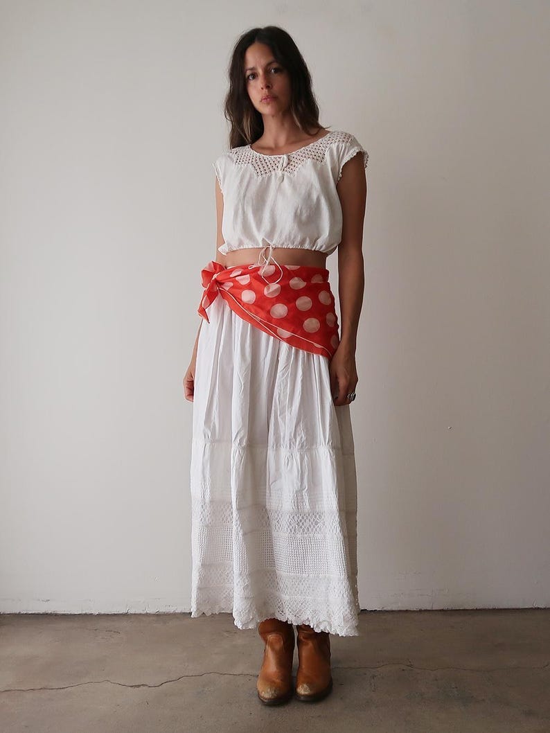 May include: A white crop top with crochet detailing and a drawstring waist is paired with a long, tiered white skirt. A red scarf with white polka dots is tied around the waist. The outfit is completed with brown leather boots.