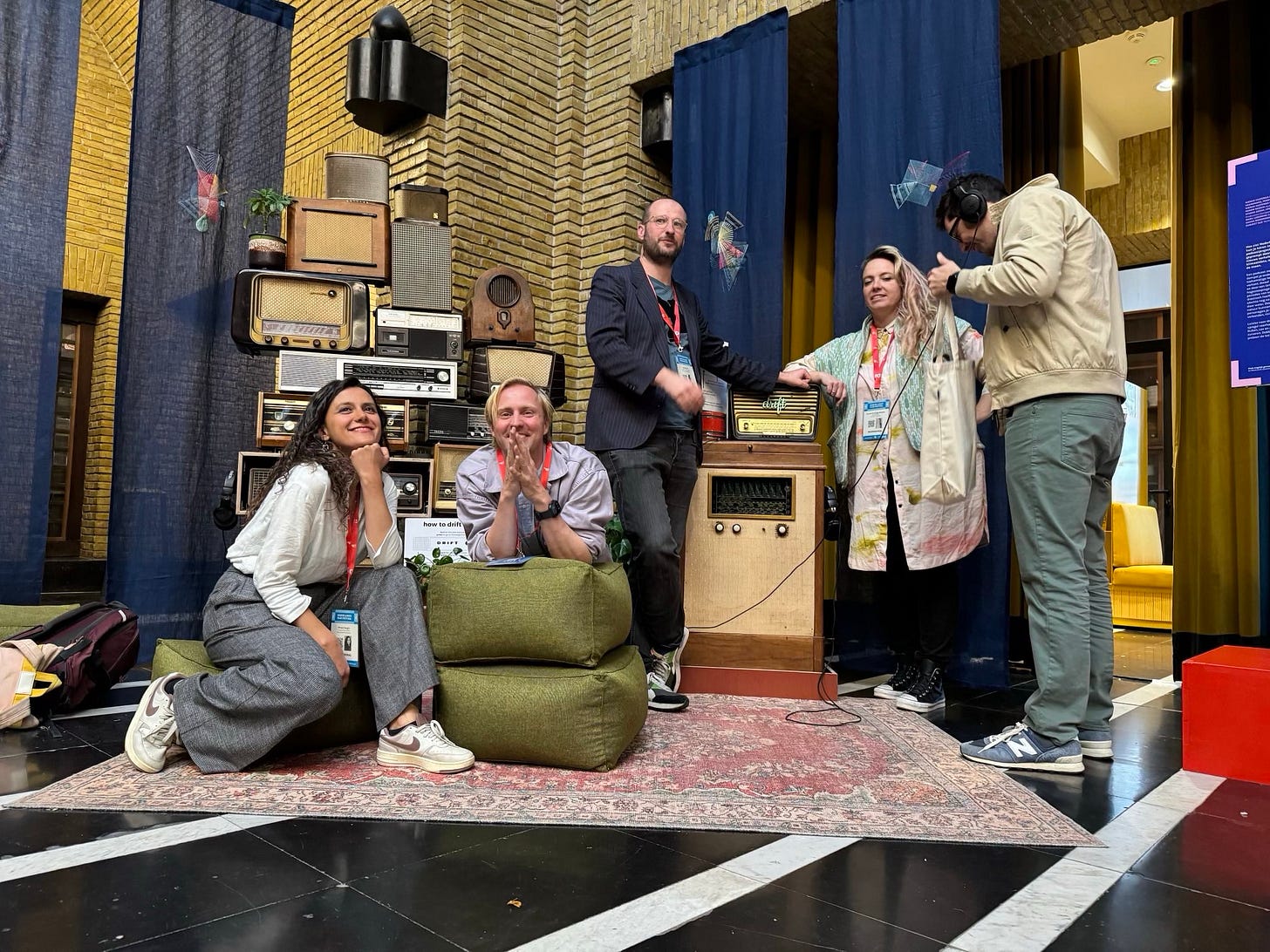 Vijf mensen poseren in een ontspannen sfeer in een decor van oude radio's in de hal van de bibliotheek op het Neude. Linksvoor zit een vrouw met lang donker haar en grijze kleding glimlachend op een groen kussen. Naast haar zit Lieven: een witte man met kort blond haar en een paarse jas. Achter hen staan drie anderen: Hay, een witte man met bril en donker pak, Nienke, een witte vrouw met lang blond haar en een lichtgekleurde jas, en een man met een koptelefoon en een beige jas. De groep lijkt heel blij in hun installatie Vijf mensen poseren in een ontspannen sfeer in een decor van oude radio's in de hal van de bibliotheek op het Neude. Linksvoor zit een vrouw met lang donker haar en grijze kleding glimlachend op een groen kussen. Naast haar zit Lieven: een witte man met kort blond haar en een paarse jas. Achter hen staan drie anderen: Hay, een witte man met bril en donker pak, Nienke, een witte vrouw met lang blond haar en een lichtgekleurde jas, en een man met een koptelefoon en een beige jas. De groep lijkt heel blij in hun installatie