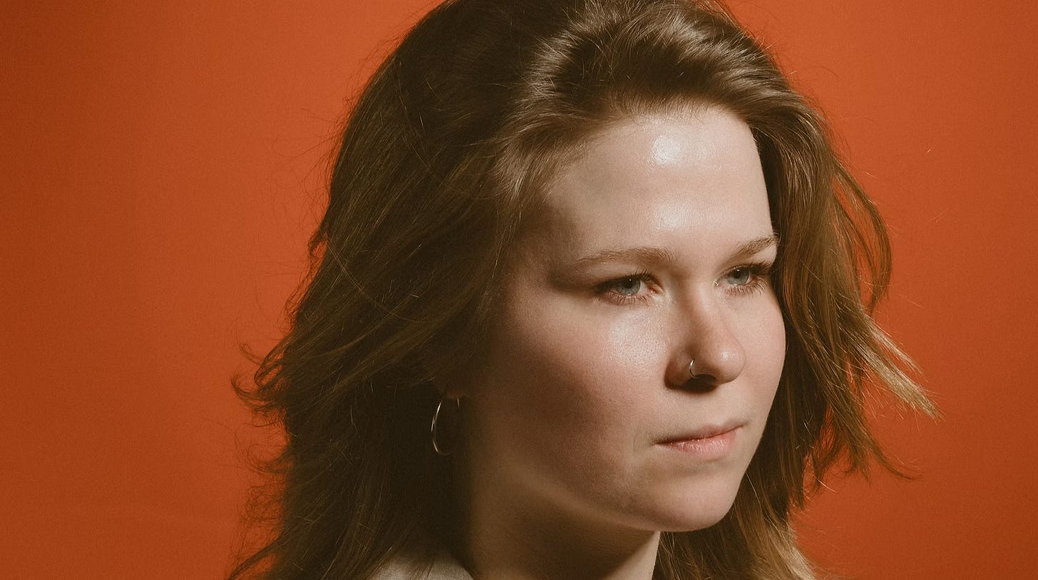 A close-up photo of Katie Spencer, in front of a vivid burnt orange background.
