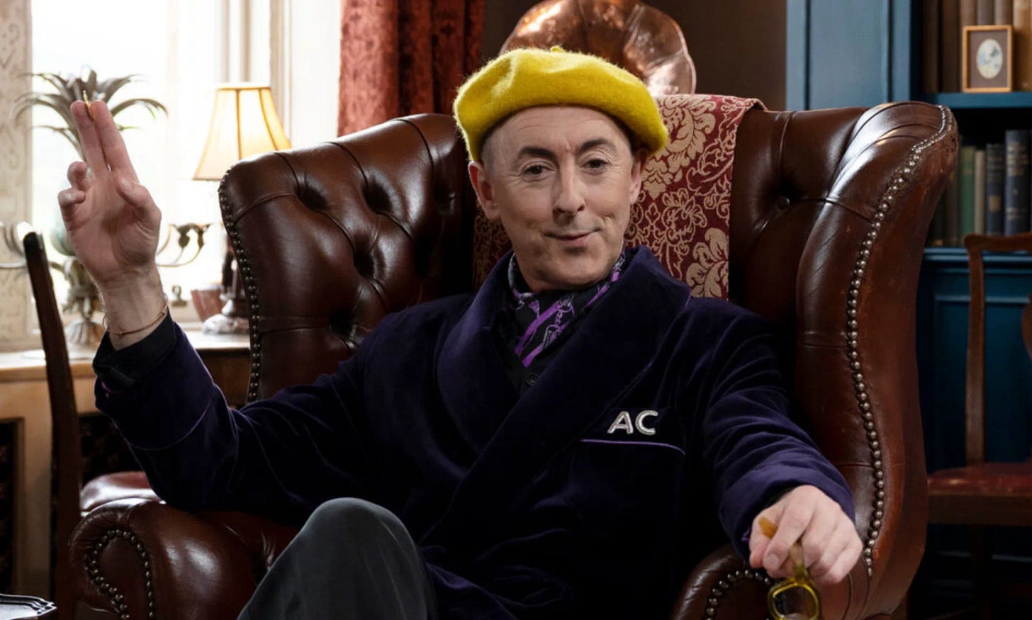 alan cumming in a chair looking sneaky