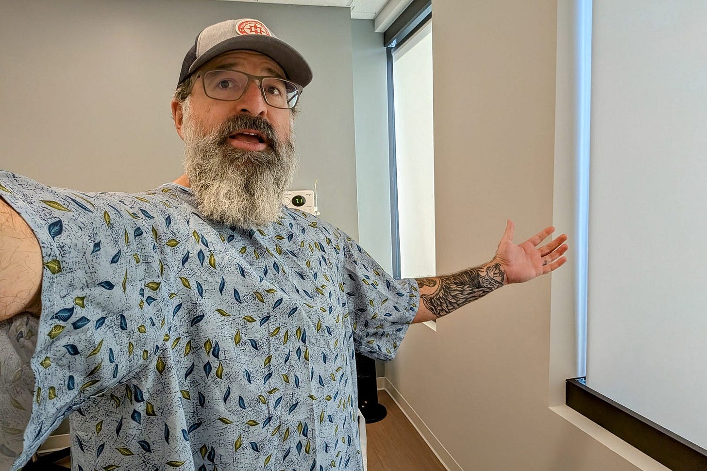 The author stands in a medical exam room wearing a hospital gown patterned with blue and green leaves. His arms are outstretched, revealing a tattooed forearm. He has a gray beard, glasses, and wears a baseball cap. A neutral wall, window, and thermostat are visible in the background, capturing a candid moment during a healthcare visit. The author stands in a medical exam room wearing a hospital gown patterned with blue and green leaves. His arms are outstretched, revealing a tattooed forearm. He has a gray beard, glasses, and wears a baseball cap. A neutral wall, window, and thermostat are visible in the background, capturing a candid moment during a healthcare visit.