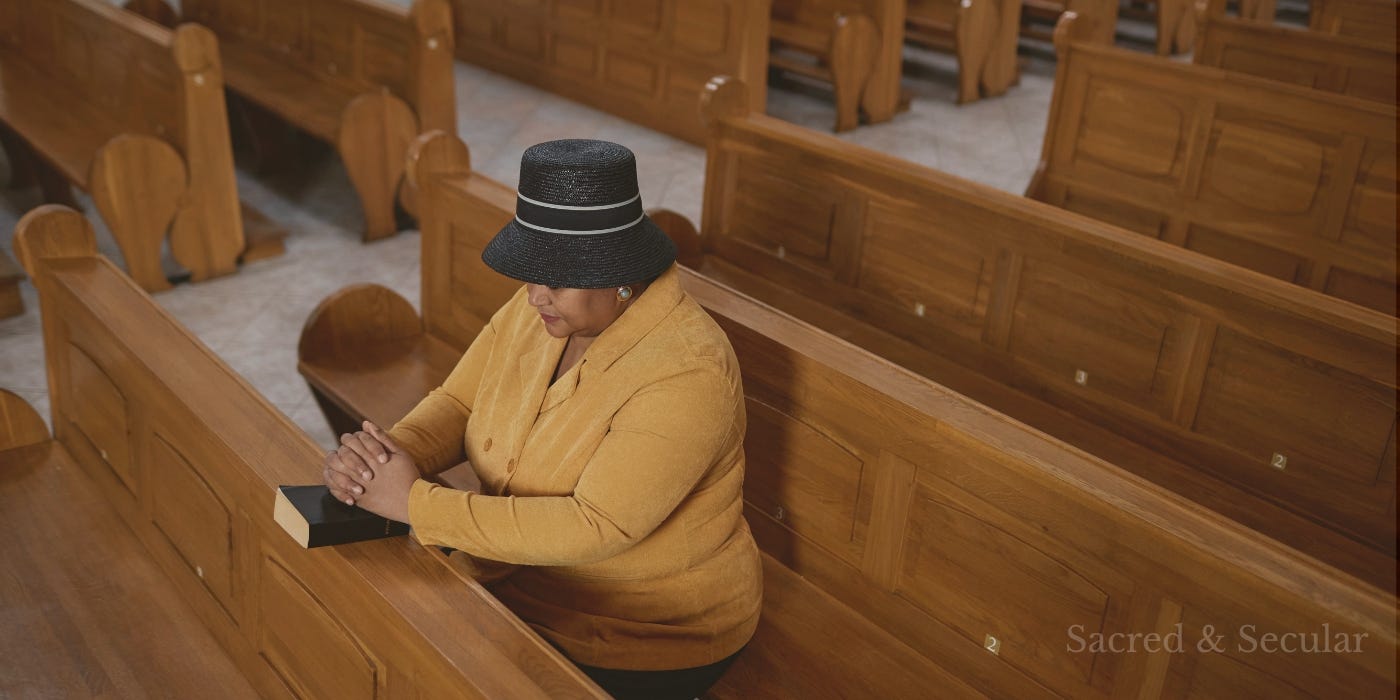 A woman in a hat sits praying in a largely empty church with rows of wooden pews.