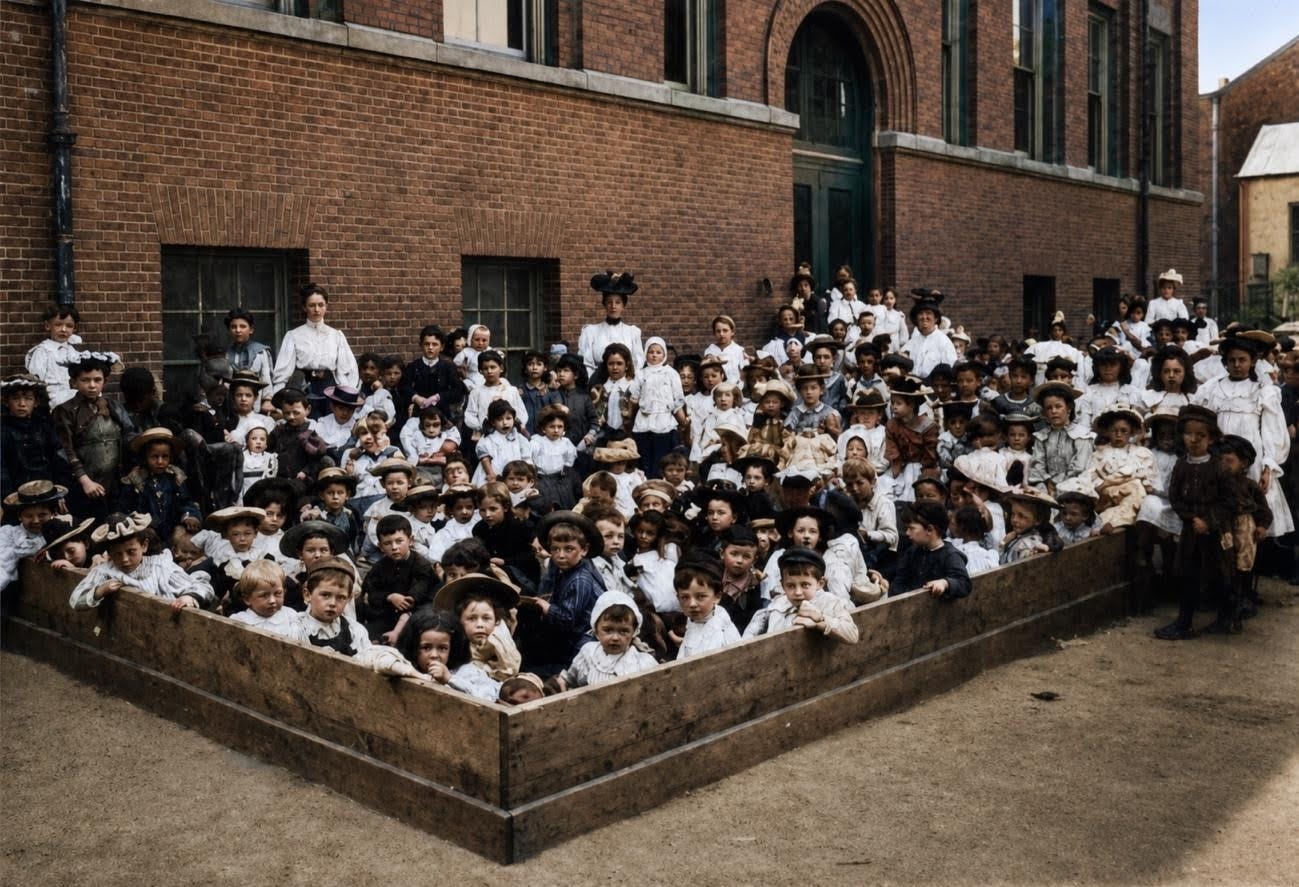 A large group of children, some in uniform, gather in and around a wooden enclosure on the playground outside a brick building for a formal group photo. A large group of children, some in uniform, gather in and around a wooden enclosure on the playground outside a brick building for a formal group photo.