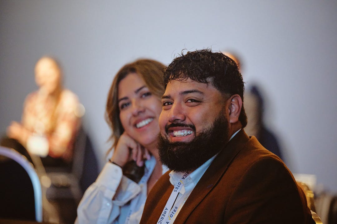 Young man in rust brown blazer grinning broadly in the audience, woman behind him smiling with her chin resting in her hand