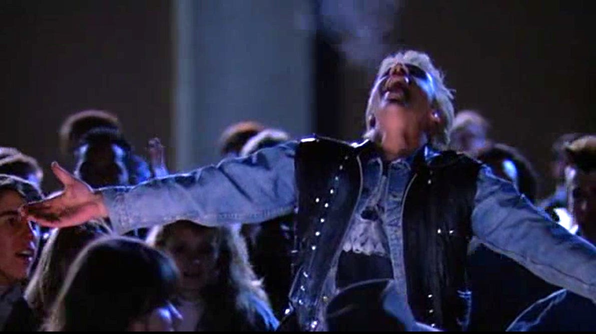 A student ringleader in denim shirt and leather jacket tosses his had back and throws his arms out in celebration.