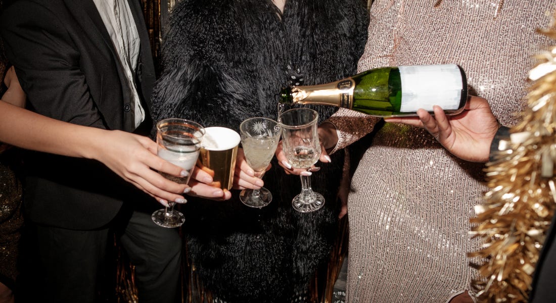 A group of people wearing black and gold clothes holding gold cups and champagne glasses, with the person to the right pouring champagne into a glass