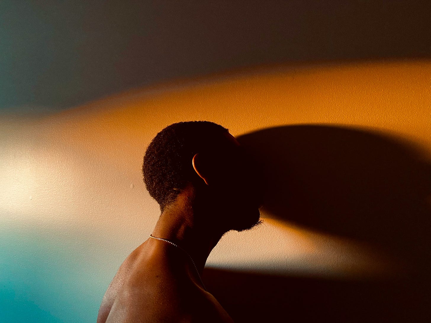 A shirtless man with short hair stands in profile against a wall, his face partly in shadow. Warm orange and cool blue light wash over the wall, creating a striking contrast that evokes reflection, longing, and emotional complexity.