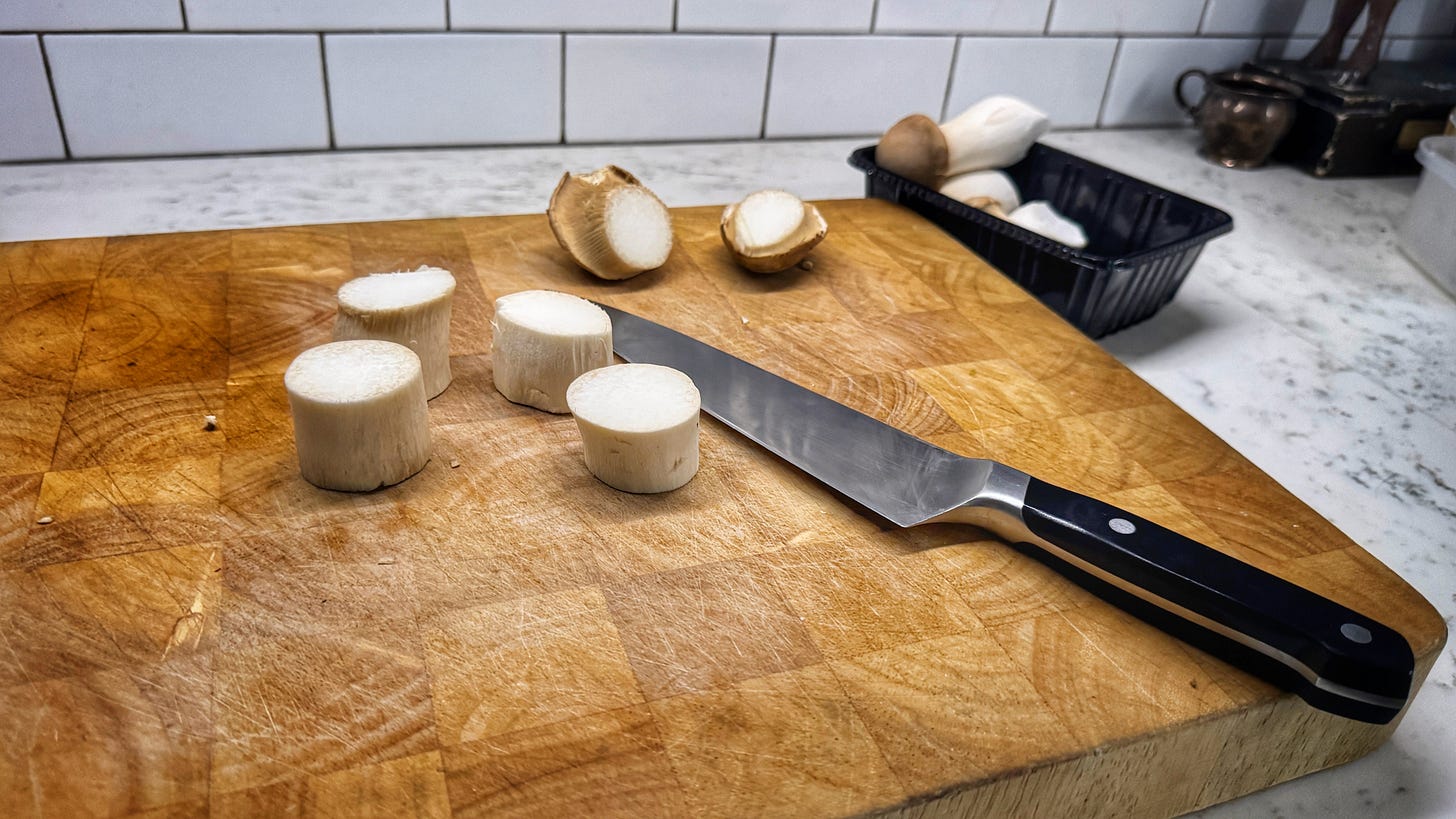 Two king oyster mushrooms on a cutting board have been cut into pieces resembling large scallops on a cutting board. Nearby are a knife, the mushrooms' caps, and more intact mushrooms in a black plastic container. Two king oyster mushrooms on a cutting board have been cut into pieces resembling large scallops on a cutting board. Nearby are a knife, the mushrooms' caps, and more intact mushrooms in a black plastic container.