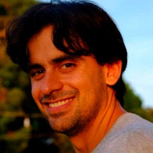 Fotografia de um homem sorrindo, com cabelos castanhos escuros, ondulados e de comprimento médio, barba curta e olhos castanhos. Ele veste uma camiseta cinza. Fotografia de um homem sorrindo, com cabelos castanhos escuros, ondulados e de comprimento médio, barba curta e olhos castanhos. Ele veste uma camiseta cinza.