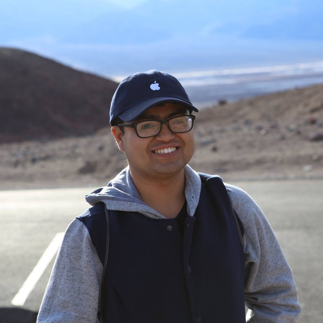 Dennis smiling wearing glasses and a navy baseball cap stands outdoors on a paved area, dressed in a gray hoodie layered under a dark vest. Rocky hills and a wide, bright sky stretch out in the background, giving the scene an open, natural feel. Dennis smiling wearing glasses and a navy baseball cap stands outdoors on a paved area, dressed in a gray hoodie layered under a dark vest. Rocky hills and a wide, bright sky stretch out in the background, giving the scene an open, natural feel.
