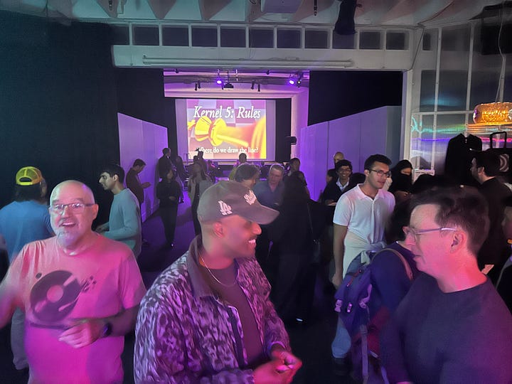 Kernel launch party in Gray Area event venue, including contributors in front of a screen, a seated audience at Jasmine's talk, magazines on a table, and various people mingling under purple lighting