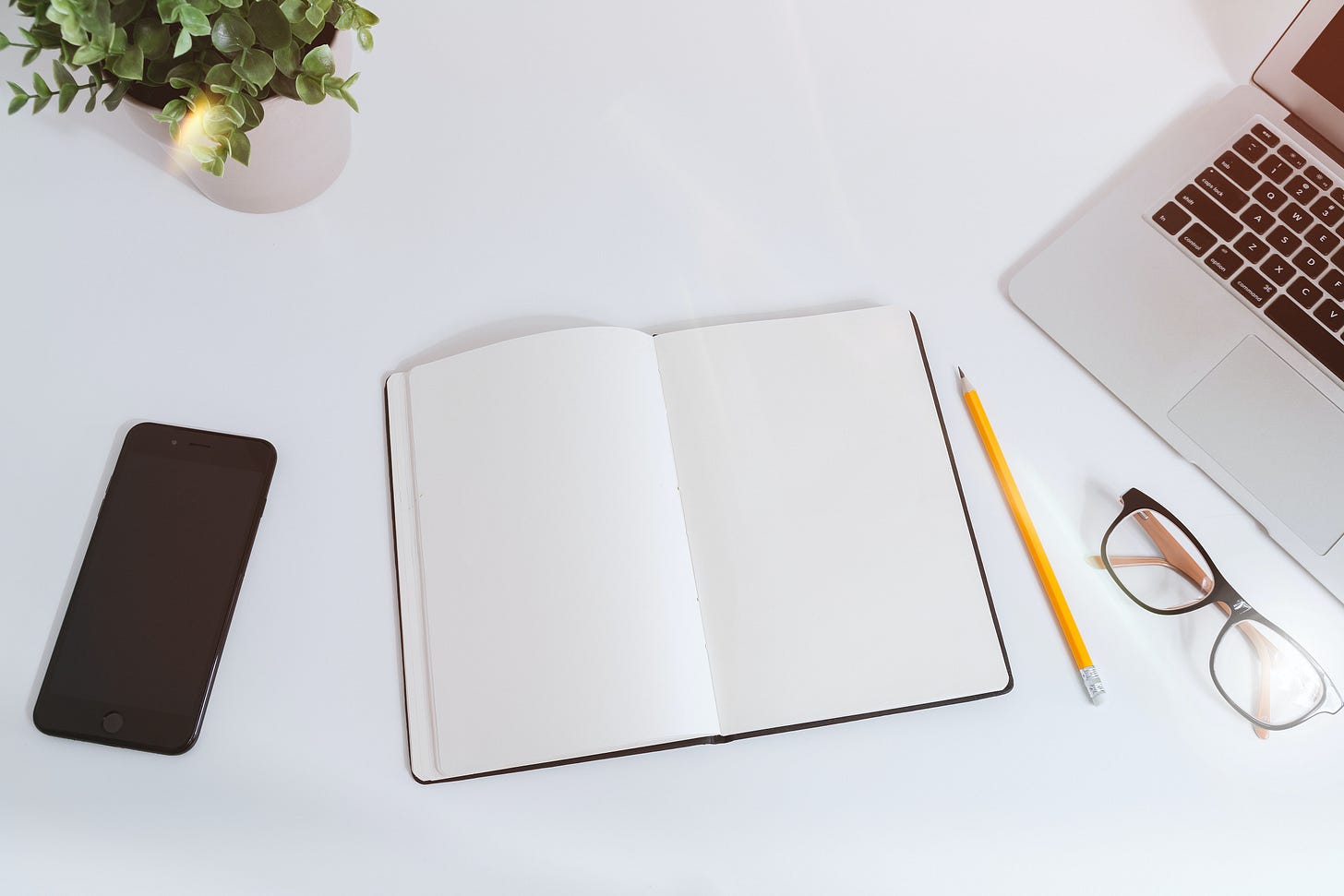 an open blank notebook, pencil, pair of glasses, phone, and laptop computer.