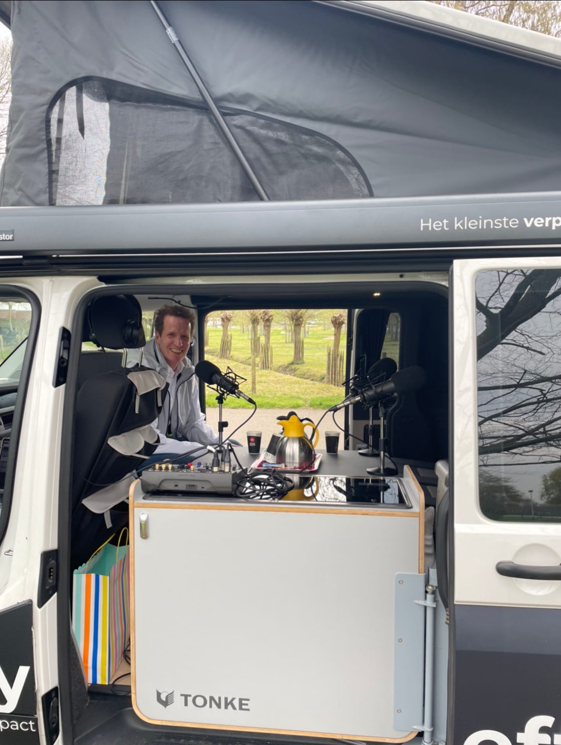 foto van een campingbusje waar microfoons in staan opgesteld, de zijdeuren zijn open en er zit een man lach achter een microfoon