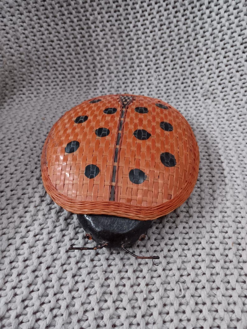 May include: A woven ladybug figurine with a black and orange body. The ladybug has black spots on its orange wings and a black head with two small black eyes.