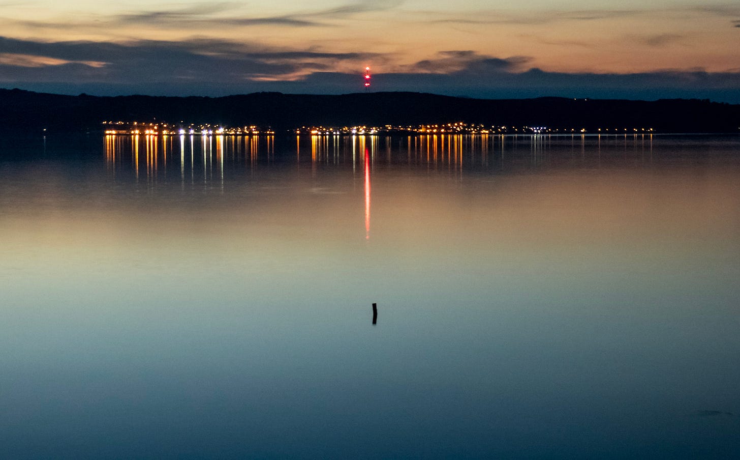 Wide dusk view across the Firth of Forth toward Edinburgh. A dark ridge and a band of city lights stretch along the far shore, reflected as long gold streaks on smooth water. A tall mast with red lights stands near center, its red reflection forming a vertical line down the water. The sky fades from warm orange near the horizon into deep blue-gray clouds, with a small dark post or buoy visible in the foreground. Wide dusk view across the Firth of Forth toward Edinburgh. A dark ridge and a band of city lights stretch along the far shore, reflected as long gold streaks on smooth water. A tall mast with red lights stands near center, its red reflection forming a vertical line down the water. The sky fades from warm orange near the horizon into deep blue-gray clouds, with a small dark post or buoy visible in the foreground.