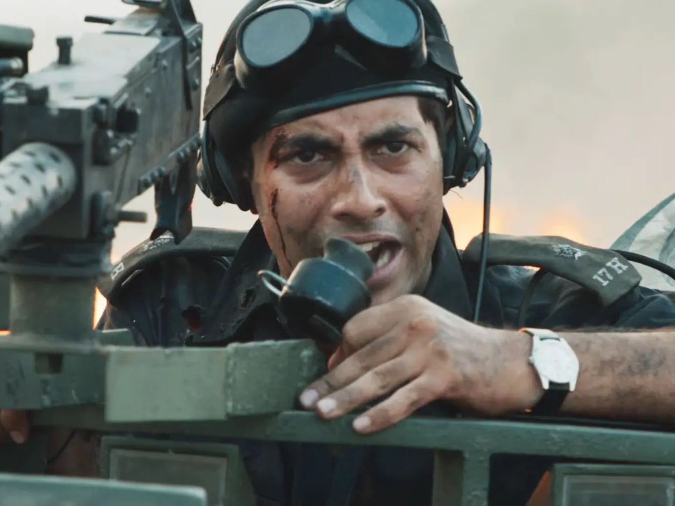 A still from Hindi film, ‘Ikkis (2026)’. Second Lieutenant Arun Khetarpal (Agastya Nanda) leans out of an army tank, wearing a helmet with goggles over his head and a radio headset, gripping a handset mic as he shouts orders. His face is streaked with dust and a cut near his temple, with a thin line of blood down his cheek; a mounted machine gun dominates the foreground, and a hazy, flame-lit battlefield glows behind him. His tense, braced posture underscore the urgency of combat.