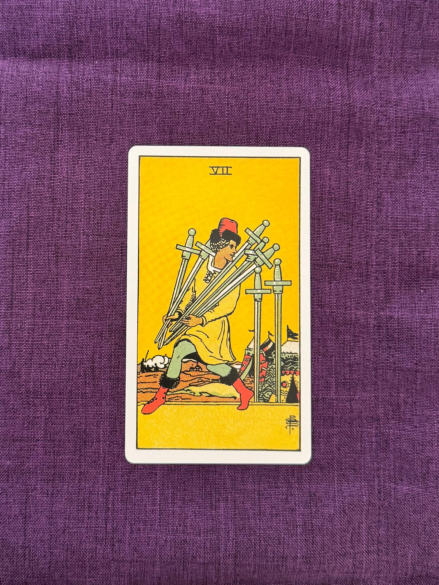 Picture of a tarot card sitting on a purple background. The card shows a man carrying five swords, while two swords remain standing on the ground.