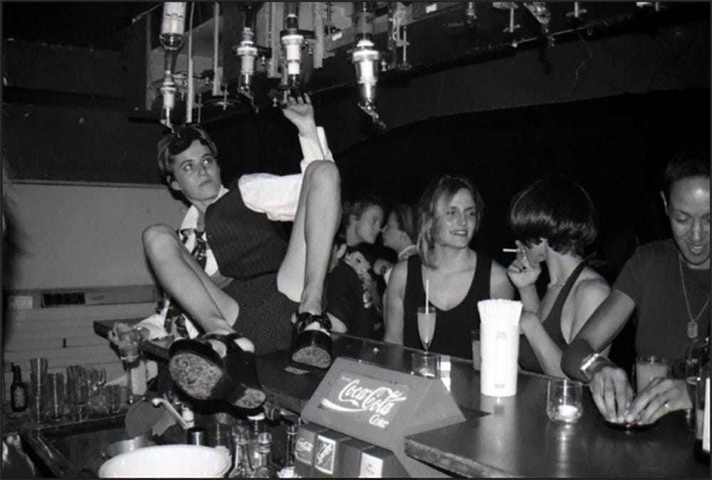 A bunch of cis lesbians are chatting and smoking against the bar counter while a short-hair cis white lesbian is sitting on the counter, legs wide open, pulling on a bottle of alcohol with her left hand, trying to fill an empty glass in her right hand.