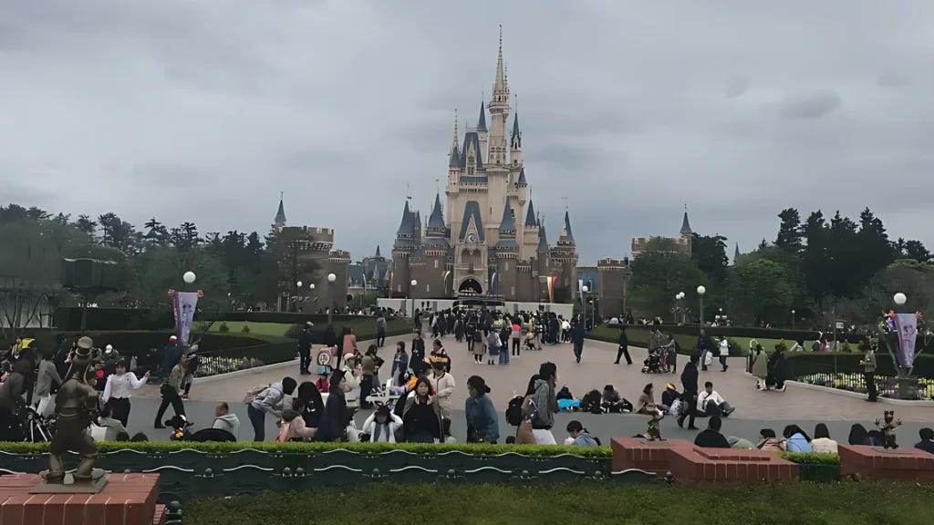 Cinderella Castle at Tokyo Disneyland under an overcast sky, seen from the crowded main plaza. Visitors mill about on the paved walkway lined with hedges and lampposts, with a costumed character statue in the foreground.