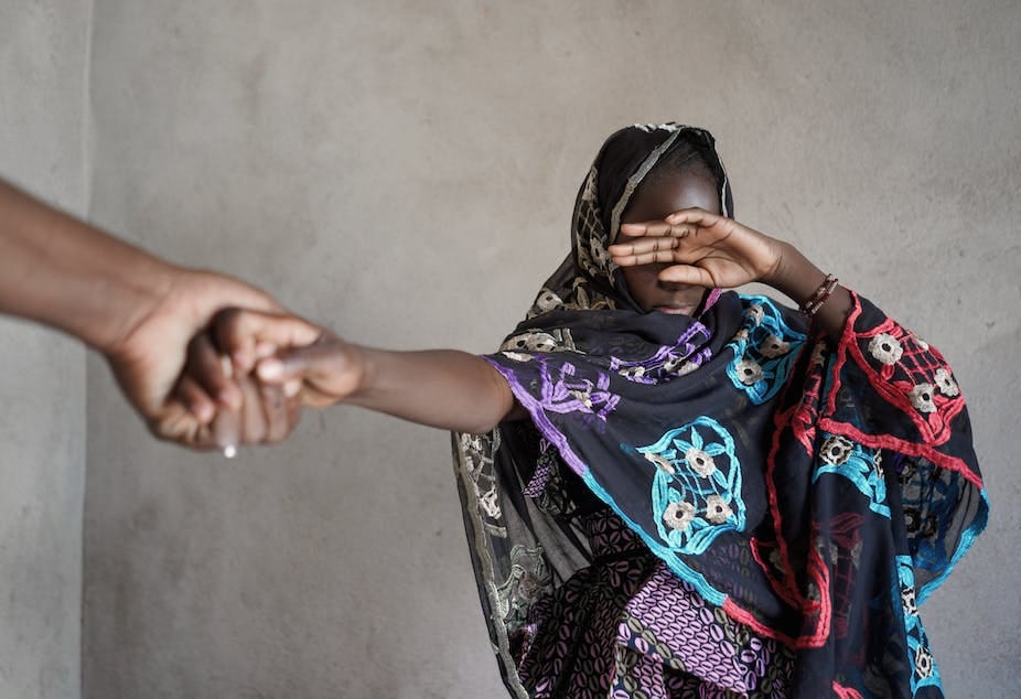 A young woman of African ethnicity wearing a black dress decorated with blue, red and purple patterns covers her eyes with one hand while an adult hand is seen pulling her by the hand