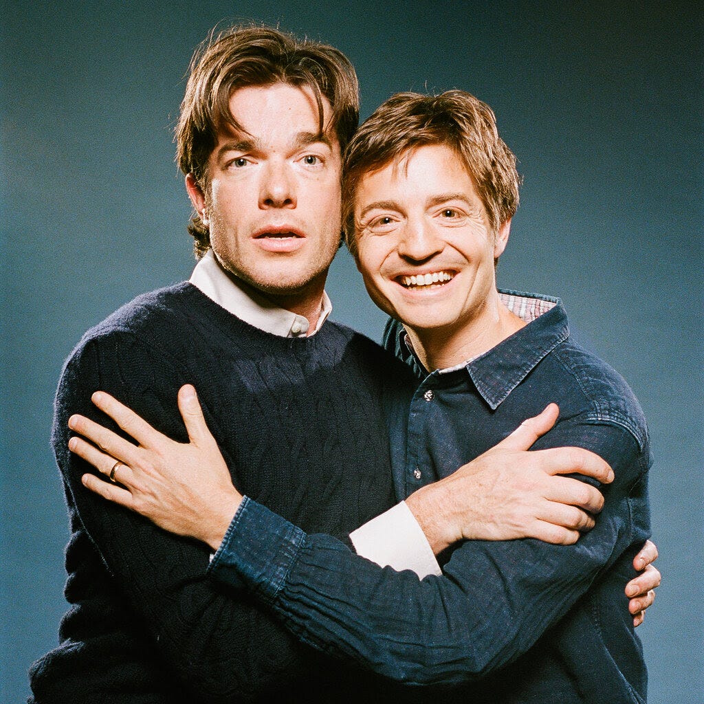 John Mulaney, in a navy blue sweater, and Simon Rich, in a button-up denim jean shirt, are sitting side by side and looking forward while embracing. 