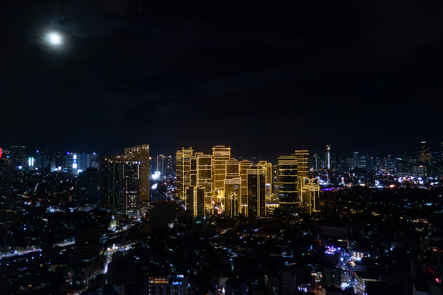 Evening photo of the Manila skyline beneath a nearly full moon. The city is mostly dark except for the busy streets lit with the headlights of cars. In the center, a collection of skyscrapers are outlined in lights