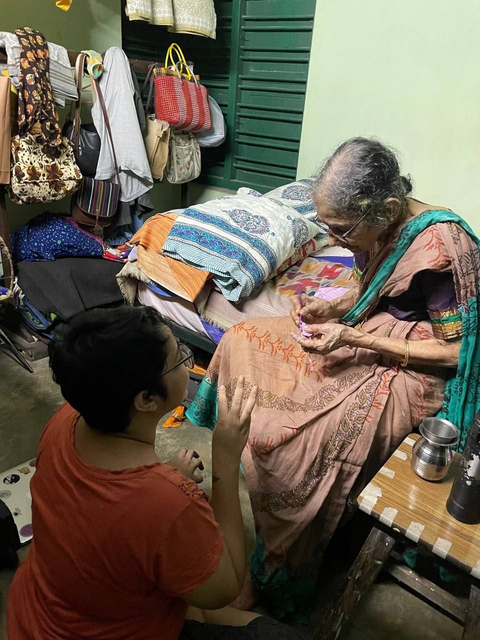 My grandmom and I seated in a room with luggage along the sides. She is trying to crochet something and I am speaking to her.