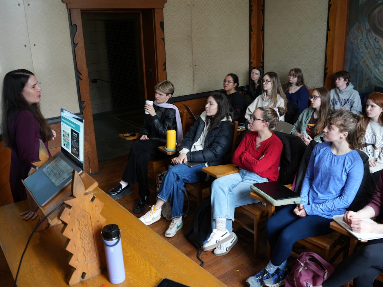 A woman stands by a wooden desk that has a computer on it. She is in a classroom. She is facing and talking to a group of students who sit in wooden desks watching her. They have notebooks and computers and look attentive. The room had wooden floors and a door frame. The walls are beige.