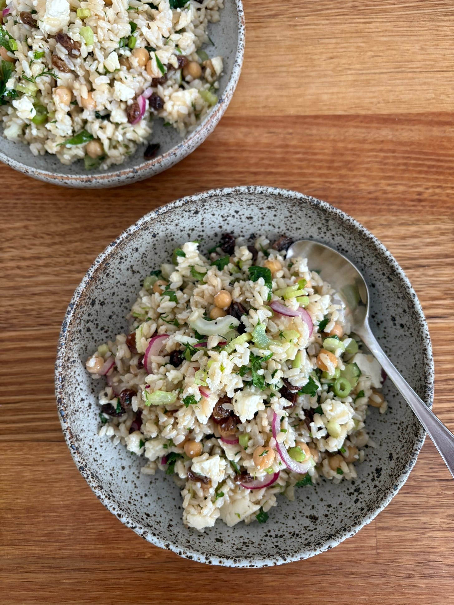 Two ceramic grey and brown speckled bowls filled with brown rice salad. The salad has red onion, sultanas, chickpeas and herbs.